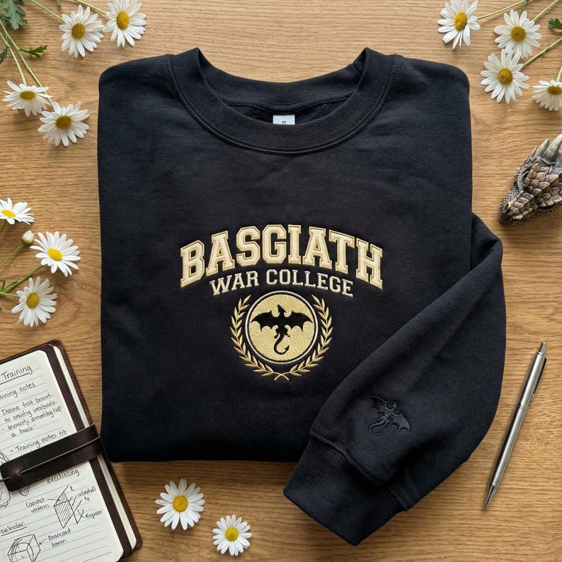 May include: Black sweatshirt with "BASGIATH WAR COLLEGE" embroidered in gold lettering. A gold dragon emblem is centered below the text. A smaller dragon emblem is on the sleeve. The sweatshirt is on a wooden surface with daisies.