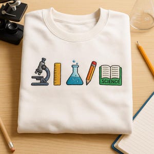 Peut inclure: Sweat-shirt crème avec des icônes brodées sur le thème de la science : un microscope, une règle, une fiole, un crayon et un livre ouvert avec le mot "SCIENCE". Le design est de style cartoon coloré.