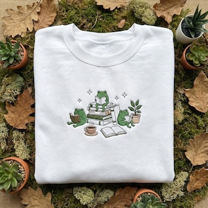 May include: White crew neck sweatshirt with an embroidered design of three green frogs reading books. One frog sits on a stack of books, others read below. A cup of coffee and a potted plant are also featured.
