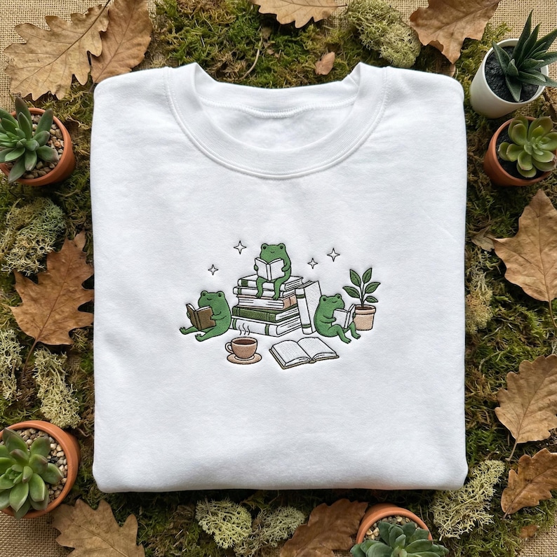 May include: White crew neck sweatshirt with an embroidered design of three green frogs reading books. One frog sits on a stack of books, others read below. A cup of coffee and a potted plant are also featured.