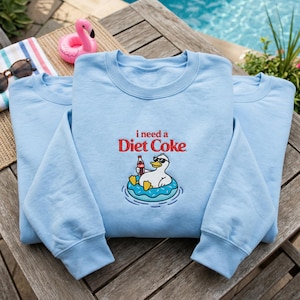 Peut inclure: Sweat-shirt bleu clair avec la phrase brodée "i need a Diet Coke". Le motif représente un canard portant des lunettes de soleil flottant sur un anneau gonflable bleu, tenant une bouteille de Diet Coke. Le sweat-shirt est présenté sur une table en bois.