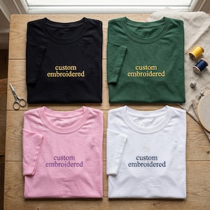 May include: Four t-shirts in black, green, pink, and white, each with "custom embroidered" in a contrasting color. The shirts are laid out on a wooden surface, with sewing supplies like scissors, thread, and an embroidery hoop visible.