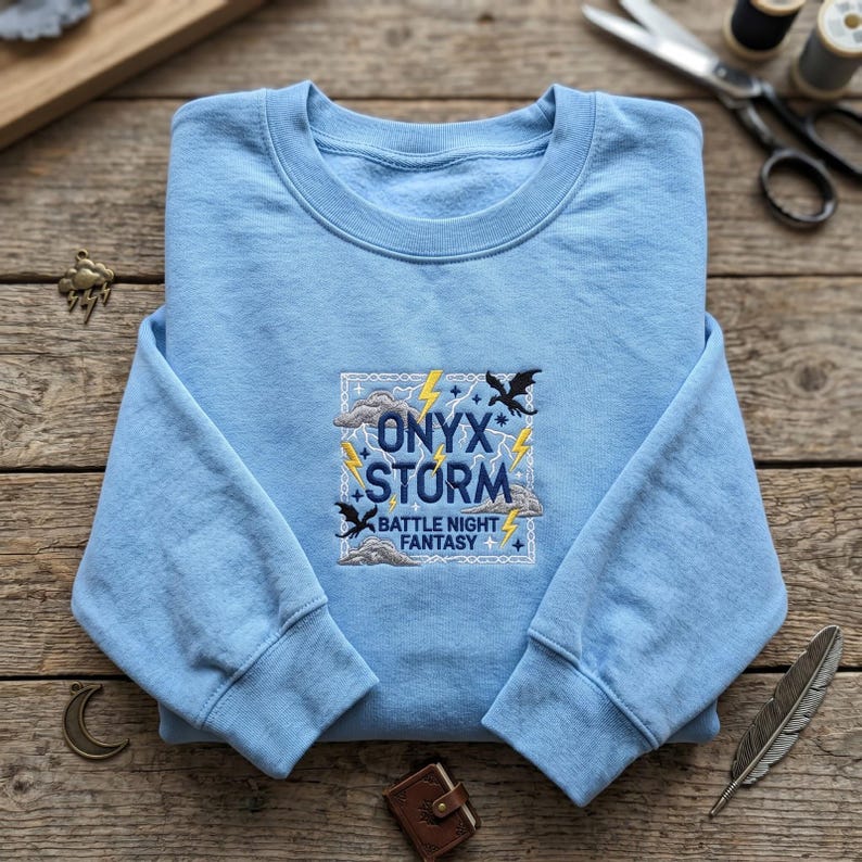 May include: A light blue crewneck sweatshirt featuring an embroidered square design on the chest. The embroidery includes the text "ONYX STORM BATTLE NIGHT FANTASY" in dark blue, surrounded by yellow lightning bolts, grey clouds, and black dragons.