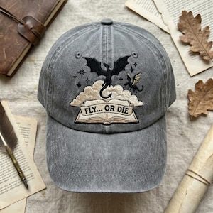 May include: Gray baseball cap featuring an embroidered design of two dragons, clouds, and stars. The text "FLY... OR DIE" is embroidered on a banner. The cap has a curved brim and a button on top.