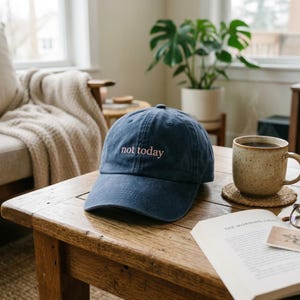 May include: A navy blue baseball cap with the phrase "not today" embroidered in pink. The hat sits on a wooden table next to a cup of coffee and an open book. The scene is set in a cozy living room.