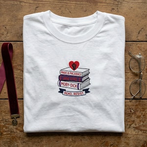 May include: White t-shirt with a stack of embroidered books featuring titles like "Pride & Prejudice" and "Moby-Dick." A red heart and the words "Read. Resist." are also embroidered on the shirt. A maroon bookmark and round glasses are on a wooden surface.