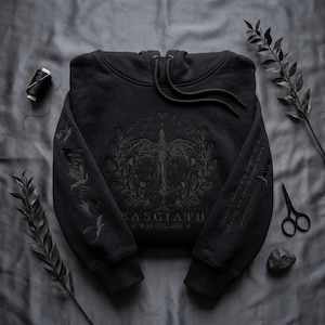 May include: Black hoodie featuring an embroidered design of a sword, laurel wreath, and the text "BASGIATH WAR COLLEGE". The sleeve has additional text. The hoodie has a drawstring hood and is made of a soft material.