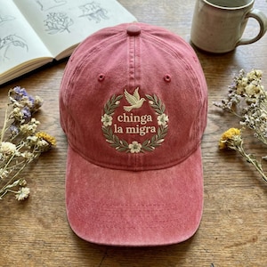May include: A faded red baseball cap with a curved peak. The cap has an embroidered design of a dove within a wreath, with the text "chinga la migra". The hat is made of a soft, worn material.