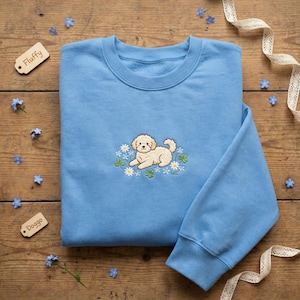 May include: A pale blue sweatshirt featuring an embroidered cream-coloured dog resting amidst white daisies and green clovers. The word "Fluffy" is on a small wooden tag. The word "Doggo" is on another wooden tag.
