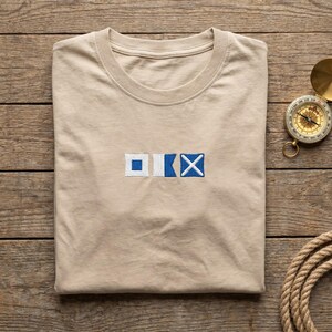 May include: A beige crew neck t-shirt featuring an embroidered design of three nautical signal flags in white and blue. The flags are arranged horizontally in the center of the chest, displaying a square, a pennant, and a cross symbol.
