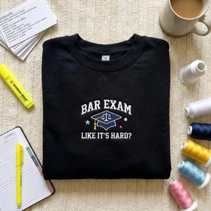 May include: Black crew neck sweatshirt with white embroidered text: "BAR EXAM LIKE IT'S HARD?". The design features a graduation cap, scales of justice, a star, and dots. The sweatshirt is displayed on a neutral-toned surface.