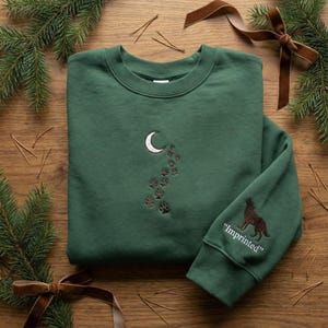 Peut inclure: Un sweat-shirt vert forêt avec une lune en croissant blanche et des empreintes de pattes brodées sur le devant. La manche présente un loup marron et le mot "Imprimé". Le sweat-shirt est présenté sur une surface en bois avec des aiguilles de pin et un ruban.