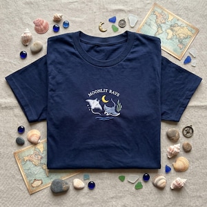 May include: Navy blue t-shirt with embroidered design featuring two manta rays, a crescent moon, and the text "Moonlit Rays." The shirt is surrounded by seashells, stones, and vintage maps, creating a nautical theme.
