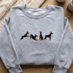 May include: A gray sweatshirt with a crew neck featuring an embroidered design of four Doberman Pinschers in various poses. The dogs are black and brown, with detailed stitching.