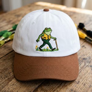 May include: White baseball cap with a brown brim and top button. The cap features an embroidered cartoon frog wearing a yellow vest and green pants, walking with a cane. The frog is set against a patch of yellow flowers.