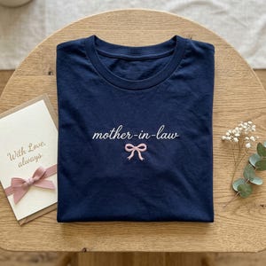 May include: Navy blue t-shirt with the embroidered text "mother-in-law" and a pink bow. A greeting card with a pink bow and the words "With Love, always" is next to the shirt. The shirt is folded on a wooden surface.