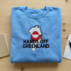 Peut inclure: Un sweat-shirt bleu clair avec une broderie représentant le Groenland, ornée du drapeau groenlandais et du texte "HANDS OFF GREENLAND" en noir. Le sweat-shirt est plié sur une surface en bois.