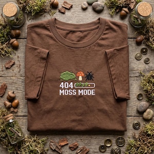 May include: A brown t-shirt featuring an embroidered graphic of a green moss patch, a red and white mushroom, and a black beetle. Below the icons, a loading bar with leaves is displayed alongside the text 404 MOSS MODE in white lettering.