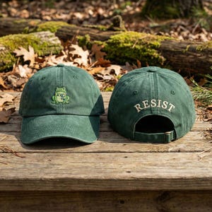 Könnte beinhalten: Zwei grüne Baseballkappen. Eine Kappe hat einen gestickten Frosch mit Schal. Die andere Kappe hat das Wort "RESIST" auf der Rückseite gestickt. Beide Kappen haben einen gebogenen Schirm und einen verstellbaren Riemen.