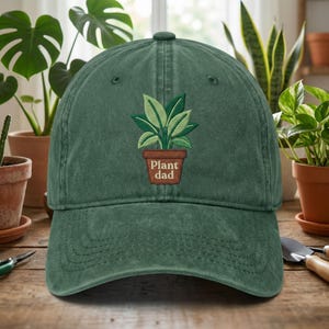 May include: A green baseball cap with a curved brim. The cap features an embroidered design of a plant in a brown pot, with the text "Plant Dad" below the plant. The cap is made of a soft, textured fabric.