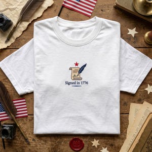 May include: A white short-sleeved t-shirt featuring an embroidered design on the chest. The graphic displays a tan parchment scroll, a dark blue quill pen, and a small red star above the text "Signed in 1776" in dark blue lettering.