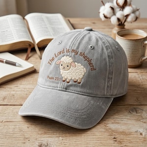 May include: Grey baseball cap featuring the embroidered words "The Lord is my shepherd" and a cartoon sheep. The text "Psalm 23:1" is below the sheep. The cap is on a wooden surface.