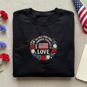 Könnte beinhalten: Schwarzes Sweatshirt mit einem gestickten Design mit einer amerikanischen Flagge und den Worten "THE ONLY THING MORE POWERFUL THAN HATE IS LOVE", umgeben von einem floralen Kranz. Das Sweatshirt ist ordentlich gefaltet.