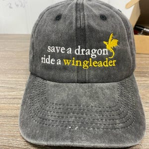 May include: A gray baseball cap with the embroidered text "save a dragon ride a wingleader" in white and yellow. A yellow dragon graphic is on the right side. The cap has a curved brim and a button on top.