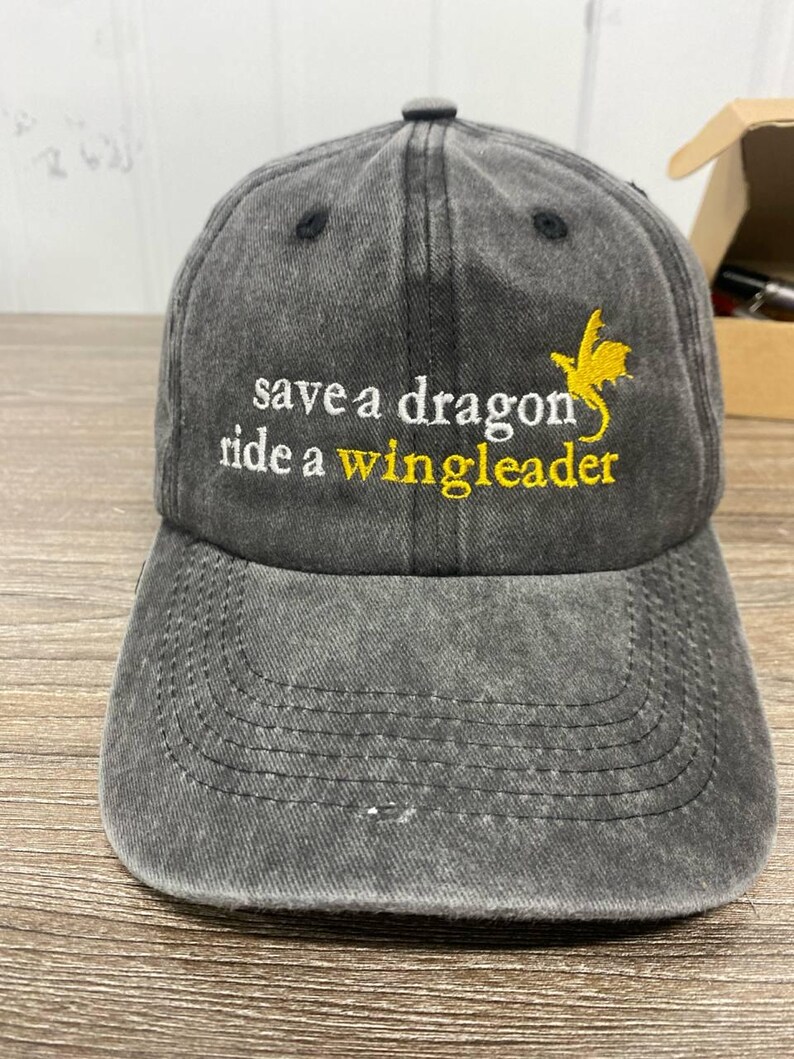 May include: A gray baseball cap with the embroidered text "save a dragon ride a wingleader" in white and yellow. A yellow dragon graphic is on the right side. The cap has a curved brim and a button on top.
