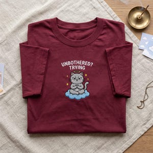Könnte beinhalten: Burgunderfarbenes T-Shirt mit der gestickten Aufschrift "UNBOTHERED? TRYING" und einer Cartoon-Katze, die auf einer Wolke meditiert. Die Katze hat einen Heiligenschein und einen grimmigen Gesichtsausdruck. Das T-Shirt liegt auf einer beigen Oberfläche.