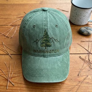 May include: A sage green baseball cap with a distressed finish, featuring an embroidered dark green pine tree graphic above the word WASHINGTON in capital letters. The cap has a structured crown, a curved brim with stitching, and eyelets.