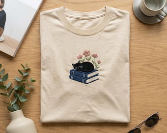 Camiseta bordada con gato durmiendo sobre libros, camiseta floral de biblioteca, regalo acogedor para lectores, bordado literario, camiseta para amantes de los gatos bibliófilos.
