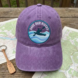 May include: A purple baseball cap with a curved peak. The cap has a circular patch with the text "SAVE OUR RIVERS" and a kayak graphic. The hat is made of a soft, textured material.