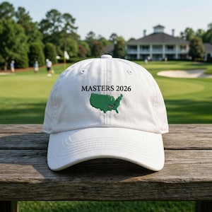 May include: A white baseball cap with the text "MASTERS 2026" embroidered in black. The cap features a green embroidered outline of the United States with a golf club. The cap is on a wooden surface with a golf course in the background.