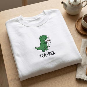Peut inclure: Sweat-shirt blanc avec un dessin de dinosaure vert tenant une tasse de thé. Le texte "TEA-REX" est imprimé en noir en dessous. Le sweat-shirt est plié sur une surface en bois clair, avec une théière visible.