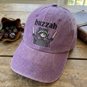 May include: A purple baseball cap featuring the embroidered word "buzzah" and a raccoon illustration holding two swords. The raccoon wears a cape. The hat is made of a soft, worn material.