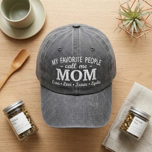 May include: A gray baseball cap with the embroidered text "MY FAVORITE PEOPLE - call me - MOM" and the names Ezra, Levi, Isaac, and Lydia. The cap is on a wooden surface with herbal tea jars and a small plant.