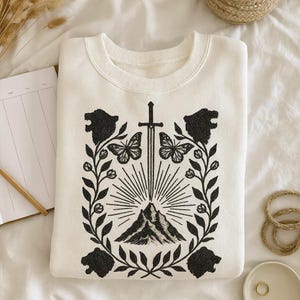 May include: Off-white sweatshirt with a black graphic. The design includes a sword, mountain, butterflies, and floral details. The sweatshirt has a crew neck and long sleeves, suitable for everyday wear.