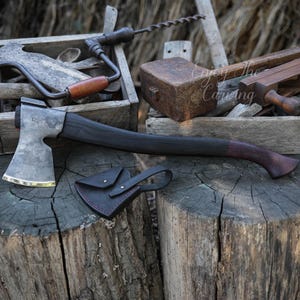 May include: A black-handled axe with a silver blade and a leather sheath. The axe is resting on a wooden stump.