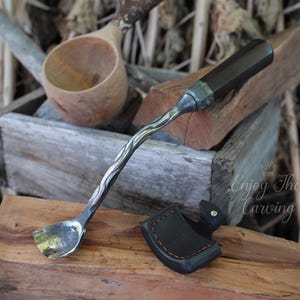 May include: A set of two wood carving tools, a curved spoon-shaped tool with a wooden handle and a small axe with a leather sheath. The tools are on a wooden surface with a wooden bowl and a piece of wood in the background. The text "Enjoy The Carving" is visible in the image.