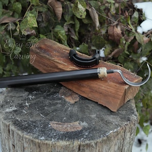 Può includere: Uno strumento di intaglio in legno nero con una lama curva e un fodero in pelle. Lo strumento è appoggiato su un pezzo di legno con il testo "Enjoy The Carving" visibile.