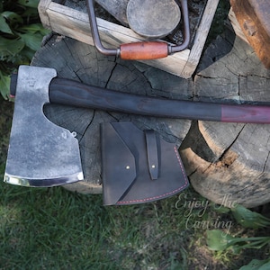 May include: A hand-forged axe with a dark wood handle and a leather sheath. The axe head is made of steel and has a polished finish. The sheath has a red stitching detail. The axe is resting on a wooden surface. "Enjoy the carving" is written on the surface.