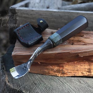 以下が含まれることがあります： ねじれたハンドルと革製の鞘が付いた黒と緑のウッドカービングツール。ツールは木の板の上に置かれています。画像には「Enjoy The Carving」というテキストが表示されています。