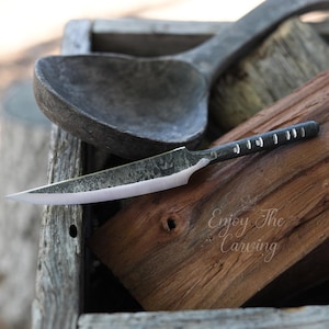 May include: A close-up of a small, hand-forged knife with a wooden handle and a textured blade. The knife is resting on a piece of wood with the text "Enjoy The Carving" printed on it.