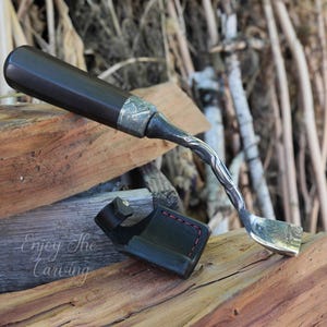 May include: A hand-forged wood carving tool with a black wooden handle and a curved blade. The tool is resting on a piece of wood and has a leather sheath.