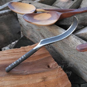 May include: A close-up of a curved, metal carving tool with a wooden handle. The tool is resting on a piece of wood with two wooden spoons in the background.