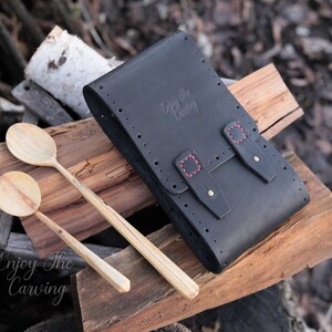 K&ouml;nnte beinhalten: Schwarze Ledertasche mit roten N&auml;hten und Messing-Akzenten. Die Tasche ist geschlossen und hat den Text "Enjoy the Carving" auf der Vorderseite gepr&auml;gt. Zwei Holzl&ouml;ffel liegen auf einer Holzoberfl&auml;che neben der Tasche.