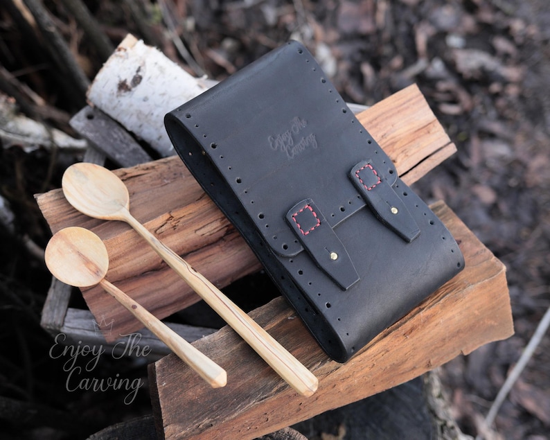 K&ouml;nnte beinhalten: Schwarze Ledertasche mit roten N&auml;hten und Messing-Akzenten. Die Tasche ist geschlossen und hat den Text "Enjoy the Carving" auf der Vorderseite gepr&auml;gt. Zwei Holzl&ouml;ffel liegen auf einer Holzoberfl&auml;che neben der Tasche.