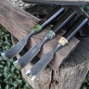 May include: Three wood carving tools with dark handles and curved blades are displayed on a wooden surface. The handles have decorative accents in shades of green and tan. The blades have a silver finish. The text "Enjoy The Carving" is visible.