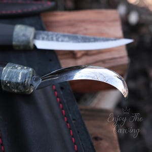 K&ouml;nnte beinhalten: Nahaufnahme eines Schnitzmessers mit schwarzem und gr&uuml;nem Griff. Das Messer ist scharf und hat eine gebogene Klinge. Das Messer liegt auf einer schwarzen Lederoberfl&auml;che.
